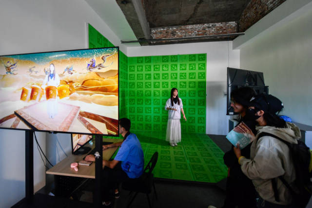 2024年7月22日，游客在敦煌莫高窟体验“寻境敦煌——数字敦煌沉浸展”后，在虚拟演播厅拍摄与洞窟虚拟场景交互的视频。“寻境敦煌”综合运用了VR虚拟现实、三维建模、游戏引擎的物理渲染和全局动态光照等新技术，高精度立体还原了莫高窟第285窟。游客戴上VR眼镜，不仅可以零距离观赏壁画、360度自由探索洞窟细节，还能“飞升”到窟顶，身临其境参与壁画故事情节，沉浸式感受敦煌文化的魅力。